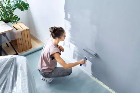 Frau malt Wand grau mit Rolle, um sie streifenfrei zu streichen. Möbel sind abgedeckt.