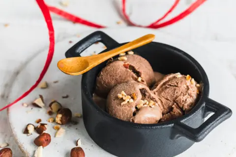Schokoladiges Nutella-Eis mit 3 Zutaten, garniert mit gehackten Haselnüssen in einer schwarzen Schale, daneben eine Holz-Löffel und rote Bänder.