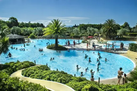 En stor pool med många badande människor, palmer och en vattenrutschkana. Perfekt för camping i Italien.