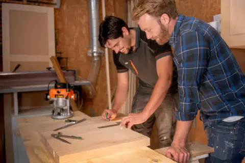Carpinteros trabajan con madera calabo en un taller. Uno marca una pieza de madera mientras el otro observa.