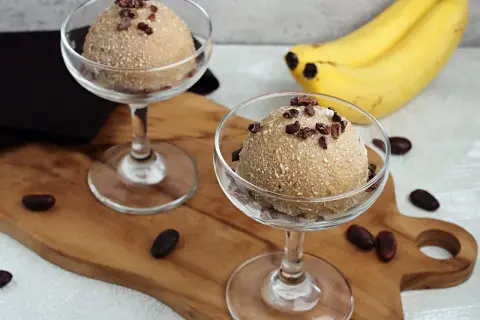 Zwei Gläser mit cremigem Apfel Zimt Eis, garniert mit Kakaonibs. Bananen im Hintergrund.
