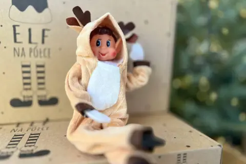 Uroczy elf w stroju renifera z uszkami i brązowymi rogami siedzi na kartonie z napisem "Elf na półce".