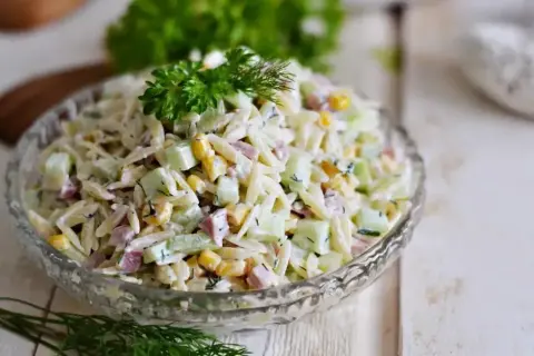 Pyszna sałatka z makaronem orzo, ogórkiem, kukurydzą i szynką, udekorowana natką pietruszki i koperkiem.