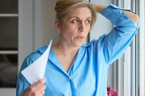 Kobieta w niebieskiej koszuli wachluje się kartką, czując uderzenia gorąca. Czy to już moment, kiedy ustępują objawy menopauzy?
