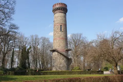 Der Toelleturm in Wuppertal, ein steinerner Aussichtsturm, ragt vor kahlen Bäumen und blauem Himmel auf. Eine Wendeltreppe führt zur Aussichtsplattform.