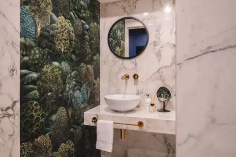 Łazienka z tapetą w leśny wzór i marmurowymi płytkami. Złote baterie i lustro tworzą elegancką aranżację.