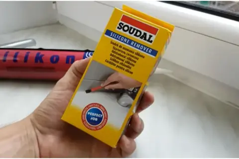 Ręka trzymająca opakowanie środka do usuwania silikonu Soudal. W tle widać tubkę z silikonem.