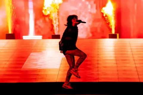 Une artiste chante sur scène, entourée de flammes spectaculaires. Cette photo de concert capture l'énergie d'un spectacle enflammé.