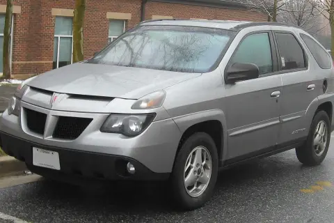 Srebrny Pontiac Aztek zaparkowany na mokrym asfalcie, z ceglanym budynkiem w tle.