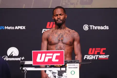 A fighter with tattoos stands on a scale, eyes closed, preparing for weigh-ins. The scale's weight brackets are visible.