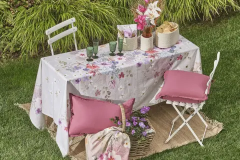 Un tavolo da giardino apparecchiato con una tovaglia floreale, bicchieri verdi, cesti e cuscini rosa. Un'idea per come rivestire un tavolo con stoffa per un picnic.