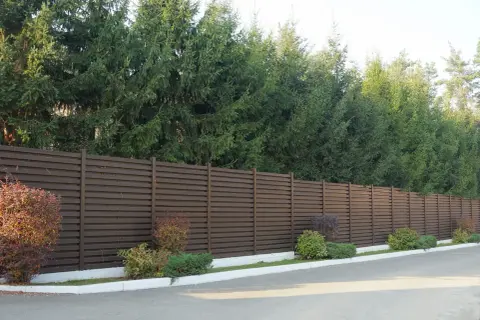 Ciemnobrązowy płot lamelowy, za którym rosną wysokie świerki, stanowi świetne rozwiązanie, czym zasłonić płot od sąsiada.