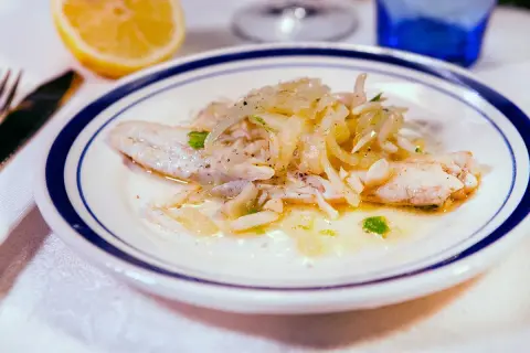 Delicato branzino in padella con cipolle e prezzemolo, servito su un piatto bianco con bordo blu. Accanto, uno spicchio di limone.