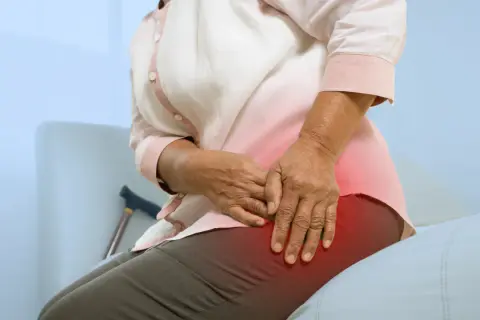 Une femme âgée se tient la hanche, souffrant de mal de hanche et sciatique. Une canne est visible derrière elle.