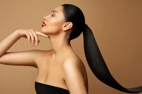 Frau mit glatten, schwarzen Haaren nach hinten gebunden, die ihr Gesicht und ihre Schultern umrahmen. Sie trägt roten Lippenstift.