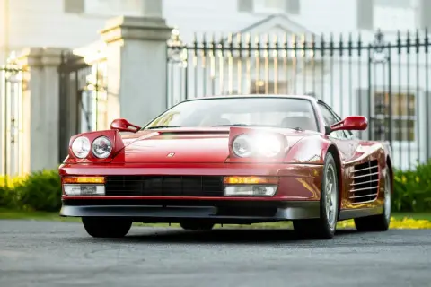 Czerwone Ferrari Testarossa z podnoszonymi światłami, zaparkowane przed elegancką posiadłością.