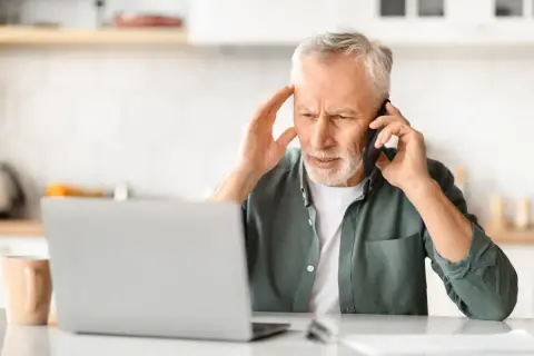 Zmartwiony mężczyzna rozmawia przez telefon, trzymając się za głowę. Obok laptopa i kubka. Wygląda na to, że jego urlop z powodu siły wyższej został zakłócony.