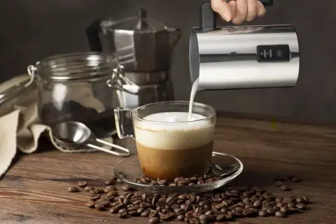 En hand häller mjölkskum i en kaffekopp, redo för ett perfekt kaffe recept. Kaffebönor sprider sig över träbordet.