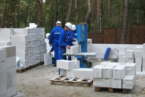 Budowa domu z bloczków betonowych. Pracownicy w niebieskich kombinezonach i białych kaskach układają materiał. Widać piłę do cięcia bloczków.