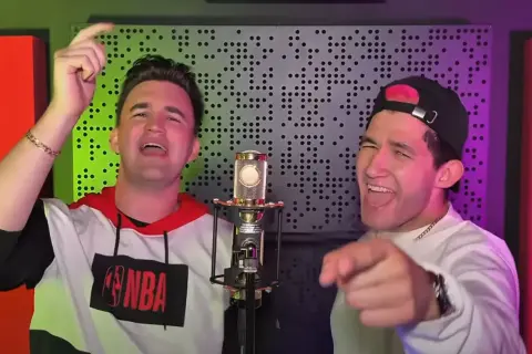 Dwóch mężczyzn śpiewa do mikrofonu. Jeden ma na sobie bluzę z logo NBA, drugi czapkę z daszkiem. W tle widać panel akustyczny.