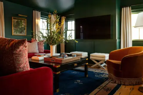A rich, jewel-toned living room with plush red sofas, an orange velvet armchair, and a dark coffee table, showcasing timeless interior design.