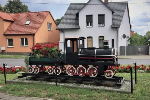 Ozdobny lokomotywa z kwiatami, jakby gotowa do odjazdu z małej stacji. W tle domy i ogródki.