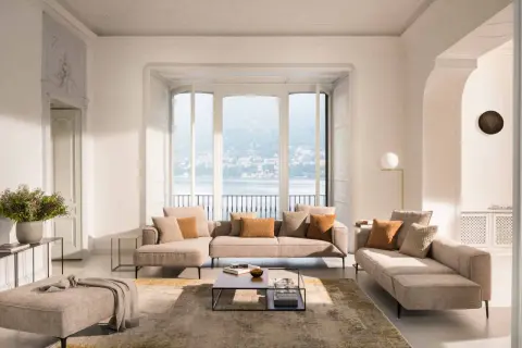 Salon lumineux avec canapé modulable et pouf dans une douce **couleur cocooning**. Vue sur un lac et des collines par de grandes fenêtres.