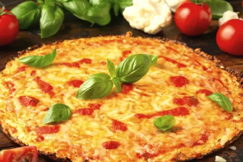 Pyszna pizza z kalafiora, z ciągnącym się serem, pomidorami i świeżą bazylią. Obok leżą pomidory i różyczki kalafiora.
