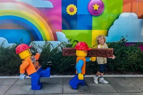 Chłopiec z uśmiechem pozuje z dwoma wielkimi ludzikami LEGO na tle kolorowego muralu. To świetne billund atrakcje dla całej rodziny!