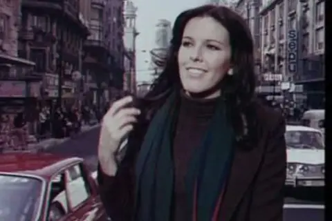 Evangelina Sobredo Galanes, con su melena oscura y bufanda, pasea por una calle urbana con edificios altos y coches antiguos.