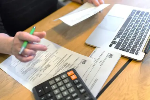 Osoba wypełnia dokumenty ZUS, obok leży kalkulator i laptop. Prawdopodobnie chodzi o zasiłek opiekuńczy.