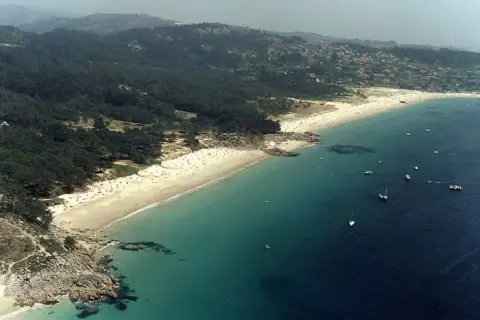 Vista aérea de la playa de Nerga, con arena dorada, aguas turquesas y barcos navegando. El paisaje se completa con pinos y colinas verdes.