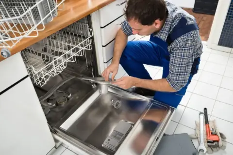 Fachowiec w niebieskim kombinezonie dokonuje podłączenia zmywarki, trzymając w rękach jej element. Obok leżą narzędzia.