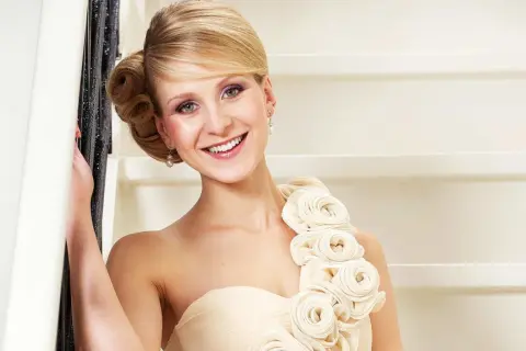 Lächelnde Frau in einem schulterfreien Kleid in Farbe ivory, verziert mit Stoffrosen. Sie lehnt an einem Geländer.