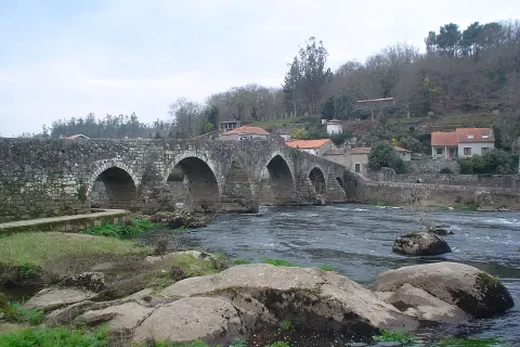 El ponte Maceira, un puente de piedra con arcos, cruza un río caudaloso. Casas con tejados rojos se asoman en la ladera.