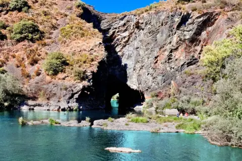 Un río de aguas turquesas fluye hacia un monte furado, una cueva natural en la roca, con vegetación y personas disfrutando del paisaje.