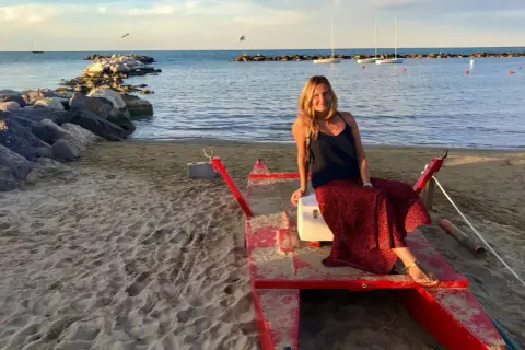 Donna sorridente su una barca rossa sulla spiaggia di Cattolica, Emilia Romagna, con il mare e i faraglioni sullo sfondo.