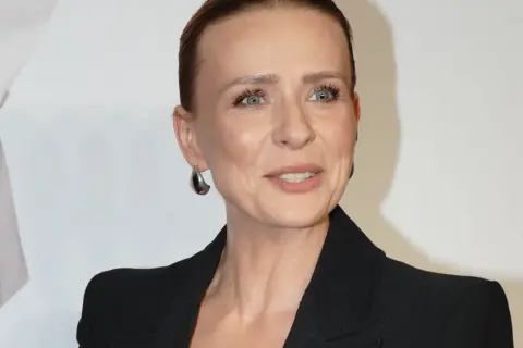 Aneta Zając w czarnej marynarce, z dużymi kolczykami, uśmiecha się. Jej partner, jeśli jest obecny, nie jest widoczny.