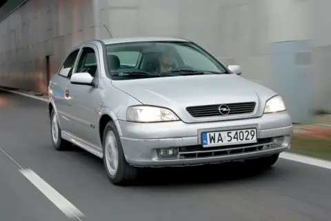 Srebrna astra 2 jedzie drogą, światła włączone, kierowca za kierownicą.