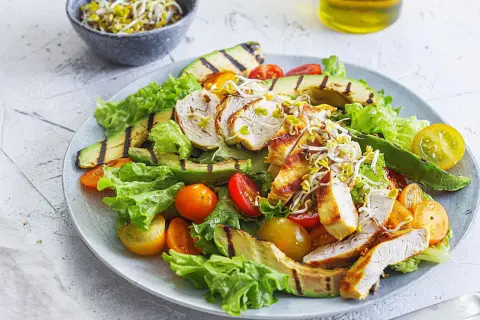 Gegrillter Hähnchensalat mit Avocado, Tomaten und Sprossen auf einem Teller.