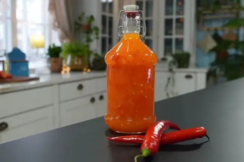 En flaska med hemmagjord sweet chili sås står på en mörk bänk, med två röda chilifrukter framför.