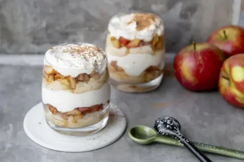 Délicieux tiramisu pomme biscuit cuillère, superposé dans des verres avec de la crème fouettée et de la cannelle. Des pommes rouges sont à côté.