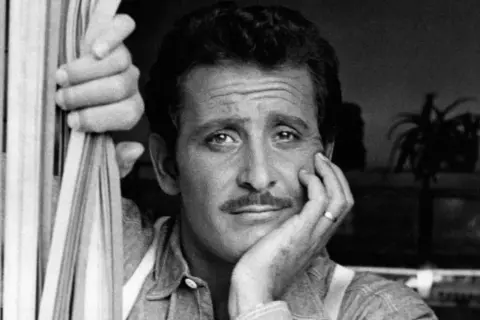 Domenico Modugno, con la mano appoggiata al viso, guarda pensieroso, mentre l'altra mano stringe delle corde.