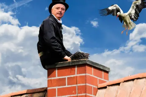 Kominiarz na dachu z miotłą, bocian w locie. PZU ubezpieczenie domu a przegląd komina to podstawa bezpieczeństwa.