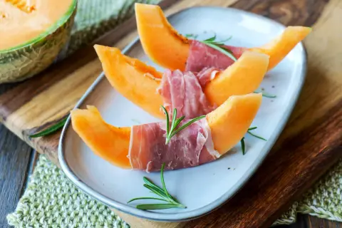 Melone mit Schinken, garniert mit Rosmarin, auf einem Teller. Ein Klassiker, der süße und salzige Aromen vereint.