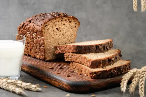 Chleb proteinowy z ziarnami słonecznika i dyni, pokrojony na kromki, obok szklanka mleka i kłosy zboża.
