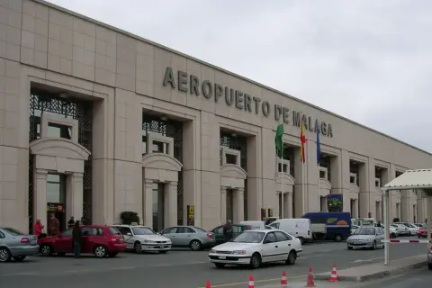 Lotnisko w Maladze, z charakterystycznym napisem na fasadzie. Przed budynkiem parkują samochody, a obok powiewają flagi.