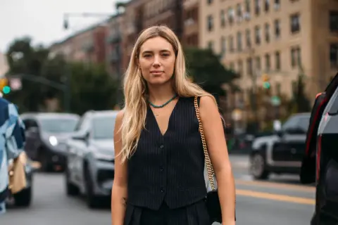 Donna con capelli biondi indossa un elegante gilet doppiopetto a righe, collana turchese e borsa a tracolla dorata, in strada.