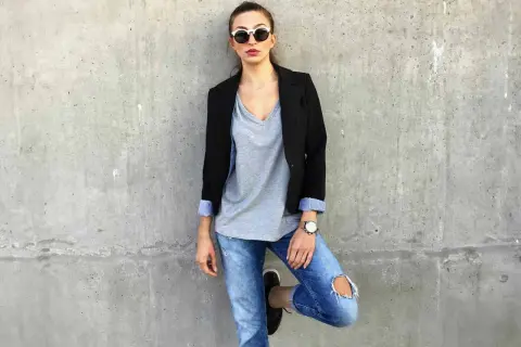 Stylisches Outfit mit Blazer: Frau in Jeans, grauem V-Shirt und Sonnenbrille lehnt an Betonwand.