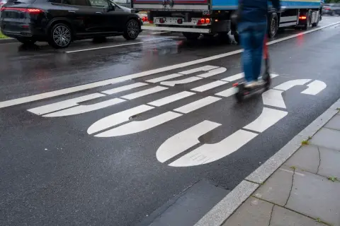 Jazda hulajnogą po buspasie grozi mandatem. Na mokrej drodze widać białe linie wyznaczające pas oraz poruszającego się po nim użytkownika hulajnogi.